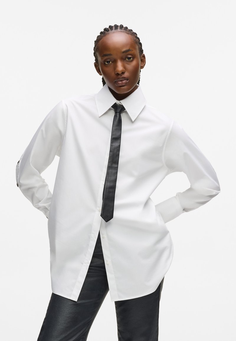 Balta garām piedurknēm krekls ar norādītu apkakli un melnu kaklasaiti, ar pogām un vaļīgu piegriezumu, valkāts ar melniem pieguļošiem biksēm.