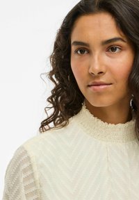 Light-colored blouse with textured chevron pattern, high frilled neckline, and sheer sleeves. Smooth fabric with subtle sheen.