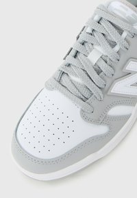 Sneaker grigio e bianco con punta perforata, accenti grigi testurizzati, lacci piatti e linguetta imbottita. Presenta un marchio prominente sul lato.