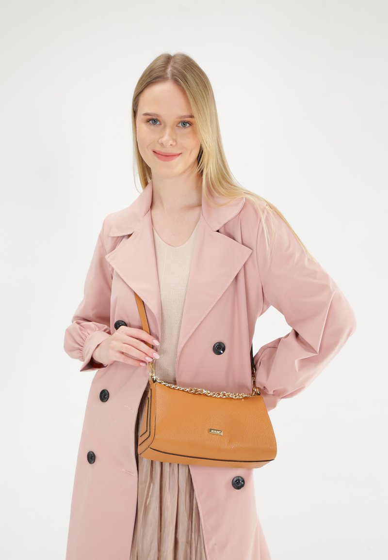 Tan læder skuldertaske med struktureret overflade, sort kant og guldkæde-rem. Modellen har en lys pink trench coat på over en nude top.