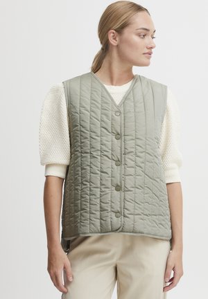 Gestepptes Weste in gedämpftem Grün mit V-Ausschnitt, Druckknopfverschluss vorne und vertikaler Naht. Getragen über einem strukturierten cremefarbenen Pullover.