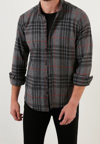 Camicia di flanella a quadri grigia con dettagli rossi, chiusura frontale con bottoni e maniche arrotolate. Il materiale sembra morbido con una finitura texture.