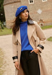 Bēša trenčkotā ar melniem akcentiem, zila vilnas džemperis un zila berete ar dekoratīvu pinumu. Melnas ādas bikses. Dabiska āra vide.