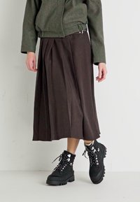 Jupe midi plissée marron associée à une veste zippée verte et des bottes noires à lacets, présentant une semelle épaisse et des détails en métal argenté.