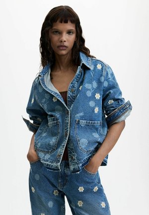 Vrouw met lang golvend haar, gekleed in een blauwe spijkerjas met bloemenpatroon en bijpassende spijkerbroek, handen in de zakken, tegen een eenvoudige lichte achtergrond.