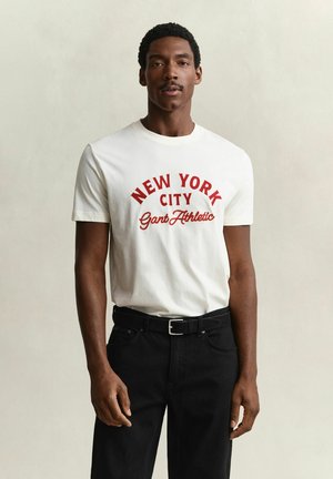 Man draagt wit T-shirt met rode tekst "New York City Gant Athletic" en zwarte broek met riem, staand tegen een effen lichte achtergrond.