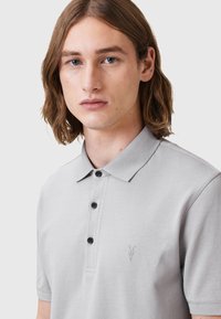 Lichtgrijze polo shirt gemaakt van gestructureerde stof, met een kraag, drie zwarte knopen en een subtiele geborduurde logo op de borst.