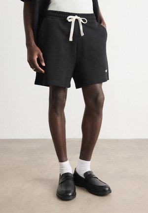 Pantaloni corti neri in cotone con cintura elastica e lacci bianchi, abbinati a calzini bianchi e mocassini neri, in piedi su un pavimento grigio chiaro.