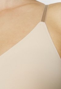 Beige behå med en slät, stretchig tyg, tunna band och en v-ringad design. Sömdetaljerna är subtila och texturen är mjuk.