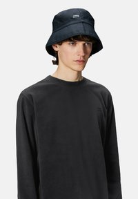 Cappello a secchiello nero realizzato in tessuto lucido con un bordo a coste. Presenta un piccolo etichetta del marchio sulla parte anteriore. Indossato con una maglietta nera a maniche lunghe.