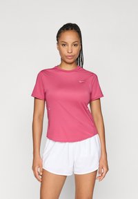 T-shirt de sport rose à manches courtes avec un col arrondi, arborant un petit logo blanc sur la poitrine et des accents discrets sur les côtés. Associé à un short blanc.