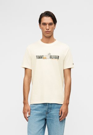 Mand iført en cremefarvet Tommy Hilfiger T-shirt med logo og grafik, kombineret med lyseblå jeans, stående mod en ensfarvet baggrund.