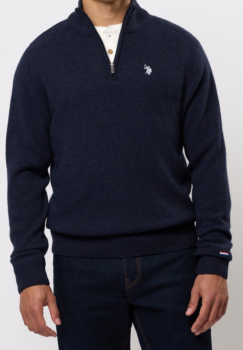 Homme portant un pull bleu marine à fermeture éclair quart, par-dessus une chemise Henley blanche, associé à un jean bleu foncé, debout devant un fond neutre.