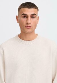 Licht beige geprofileerde trui met een ribbelkraag en lange mouwen, gedragen door een persoon met kort haar, tegen een effen achtergrond.