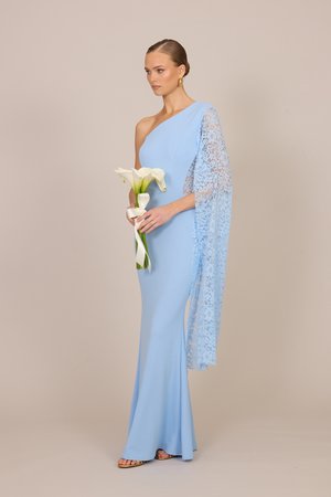 Kobieta w jasnoniebieskiej sukni na jedno ramię z koronkowym rękawem trzyma bukiet białych lilii calla na beżowym tle.