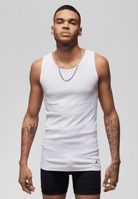 White cotton tank top with a slim fit, featuring a small black logo. Model displays noticeable tattoos. Black shorts complete the outfit.