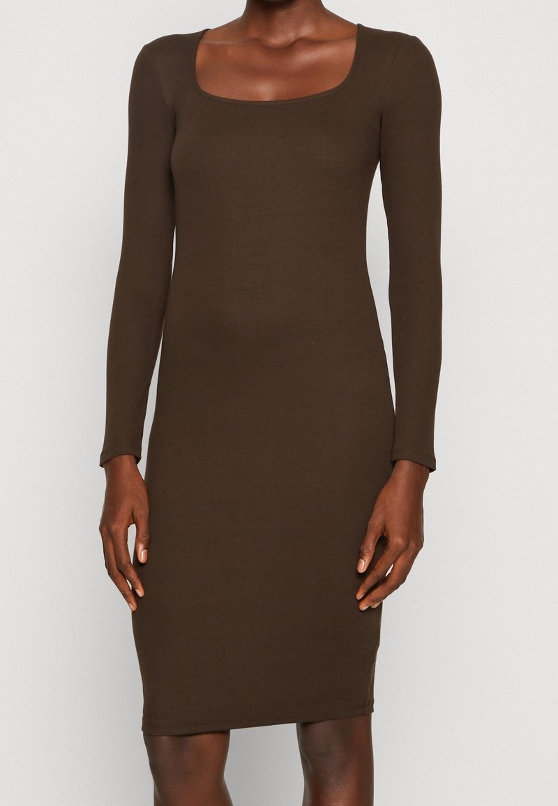 Femme portant une robe ajustée, manches longues, longueur genou, de couleur marron foncé avec un large décolleté arrondi, debout devant un fond uni.