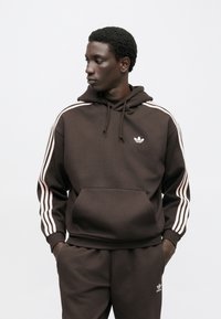 Camisola com capuz castanha com bolso frontal e detalhes brancos de três riscas nas mangas. Apresenta um pequeno logótipo branco no peito. Tecido macio e texturizado.