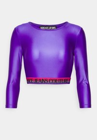 Purple cropped long-sleeve top made of glossy material, featuring a black and pink elastic waistband with "VERSACE JEANS COUTURE" printed.