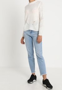 Maglione bianco a maglia con scollo rotondo, caratterizzato da dettagli a maglia aperta, abbinato a jeans in denim azzurro chiaro e sneakers nere con accenti bianchi.
