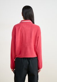 Punane fliispullover, millel on mock neck ja kontrastse heledalt roosa äär. Omab lühendatud kujundust, pikki varrukaid ja nööriga sera detaili.