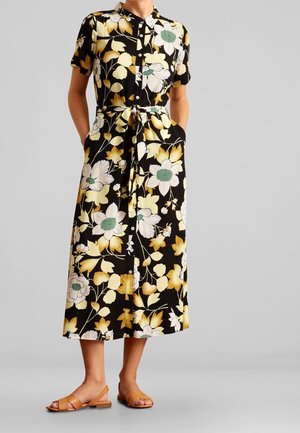 Frau trägt schwarzes Midikleid mit gelbem und weißem Blumenmuster, geschnürtem Taillengürtel und beigen flachen Sandalen, steht mit den Händen in den Taschen.