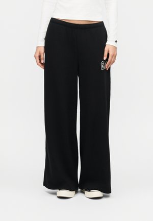 Person wearing black wide-leg pants with white "GAP" logo on thigh, white long-sleeve top, and white sneakers standing against plain background.