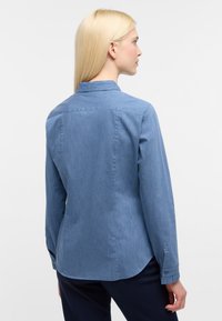 Camicia in denim blu con colletto, maniche lunghe e orlo curvato. Il materiale ha una leggera texture e presenta polsini con bottoni.