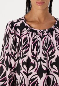 Blouse à manches courtes en tissu léger, présentant un motif floral noir et rose sur fond rose clair, avec un col rond.