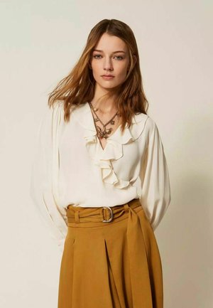 Donna con capelli castano chiaro che indossa una camicetta bianca con volant, collane a strati e una gonna senape a vita alta con una cintura larga.