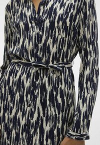 Abito camicia a maniche lunghe in un motivo astratto blu navy e crema, con una chiusura frontale a bottoni e una cintura da annodare per regolare la forma.