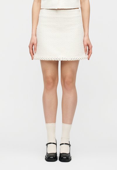 Femme portant une mini-jupe blanche texturée avec un ourlet décoratif, des chaussettes crème et des chaussures Mary Jane noires, debout devant un fond blanc.