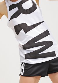 Biały top na ramiączkach z dużym czarnym napisem graficznym, w połączeniu z czarnymi szortami, które mają trzy białe paski po bokach.