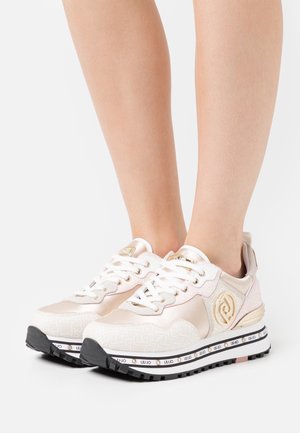Lichtroze en witte plateau sneakers met gouden logo-accenten, zwarte zolen met profiel en witte veters, gedragen op blote voeten.