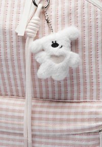Porte-clés en peluche blanche en forme d'ours sur un sac en tissu à rayures roses et beige. L'ours a un détail en forme de cœur brillant et des traits brodés noirs.
