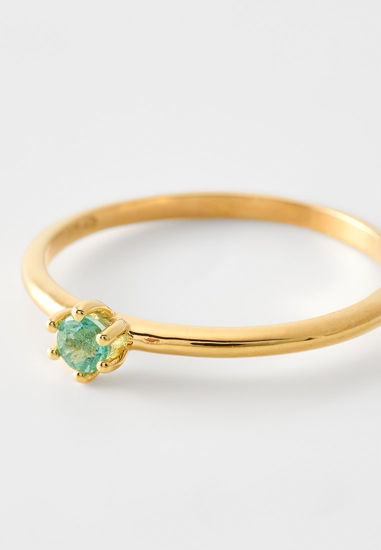 Goldring mit einem kleinen runden grünen Edelstein, der von sechs Krappen gehalten wird, auf einem weißen Hintergrund.