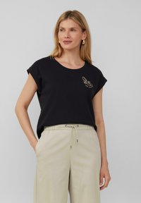 Zwarte katoenen t-shirt met korte mouwen, voorzien van een gouden geborduurd bijenontwerp op de linkerborst, gecombineerd met lichtbeige broek met trekkoord.