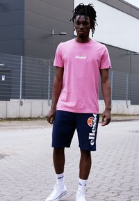 Rosa Baumwoll-T-Shirt mit "ellesse"-Logo, kombiniert mit marineblauen Shorts mit Logodetails. Weiße Socken und Sneaker runden das Outfit ab.