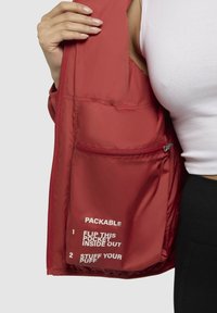 Red packable jacket with a smooth nylon interior, featuring a zippered pocket and white text instructions on storage.