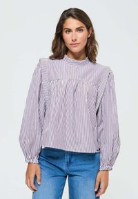 Femme portant un chemisier à manches longues rayé verticalement en violet et blanc avec des épaules volantes, et un jeans bleu, se tenant devant un fond blanc.