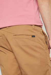 Brązowe szorty z płóciennego materiału z pochyloną kieszenią, z czarną naszywką logo Volcom, zestawione z różową koszulą z krótkim rękawem.