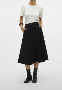 Jupe midi noire avec des plis et des poches, portée avec un haut blanc à manches courtes. Complétée par des bottes à talons noires. Fond uni.