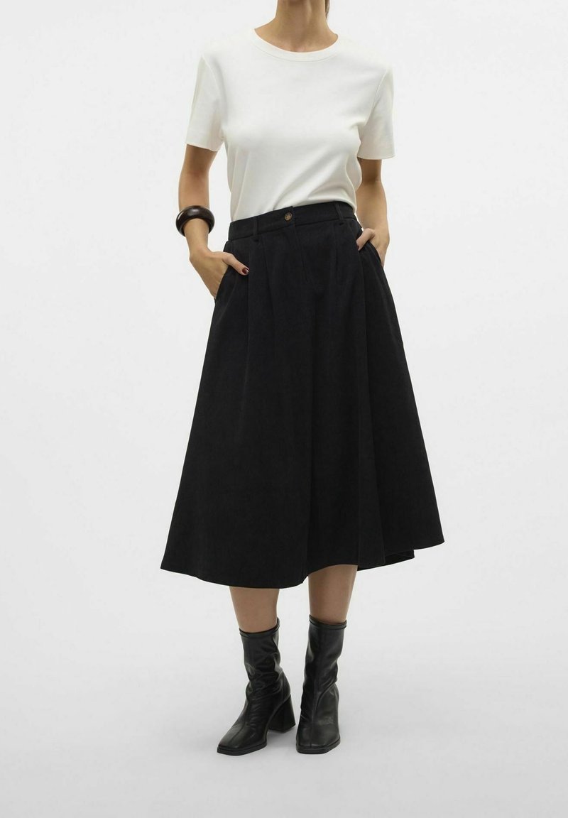 Jupe midi noire avec des plis et des poches, portée avec un haut blanc à manches courtes. Complétée par des bottes à talons noires. Fond uni.