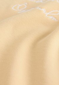 Žlutá látka s hladkým povrchem, se vzorem bíle vyšívaného textu. Materiál vypadá lehce a má mírný lesk.
