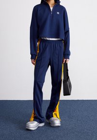 Sudadera recortada con cremallera en color navy y logo blanco, combinada con pantalones navy que tienen franjas amarillas laterales. El modelo lleva puestos unos zapatos plateados gruesos y una bolsa negra.