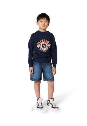 Sweat-shirt bleu marine avec "VOLTAIRE" imprimé en orange, associé à un short en denim bleu. Baskets montantes noires et blanches avec des lacets lâches.