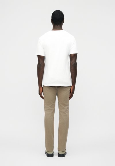 Homme debout de dos portant un t-shirt blanc uni à manches courtes, un pantalon beige et des chaussures noires sur un fond blanc.