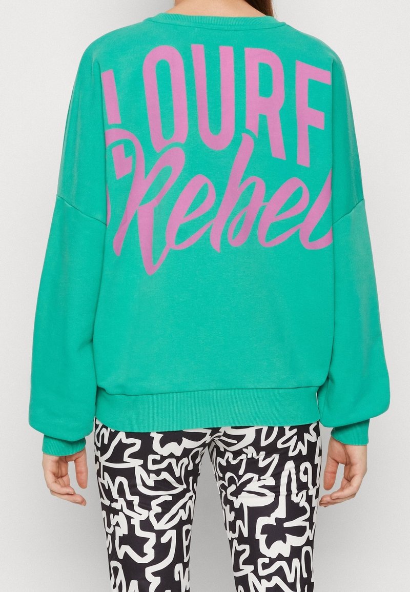 Personne portant un sweat-shirt turquoise avec un grand texte rose "Surf Rebel" dans le dos et un pantalon à motif abstrait noir et blanc, debout devant un fond uni.