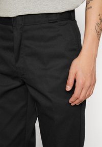 Black fabric trousers with a smooth texture, featuring a straight cut and a waistband. A hand rests on the leg, showing natural nails.