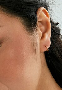 Un orecchino a cerchio dorato si trova nel lobo, con un design liscio e arrotondato. L'orecchio ha un tono di pelle naturale e capelli scuri.